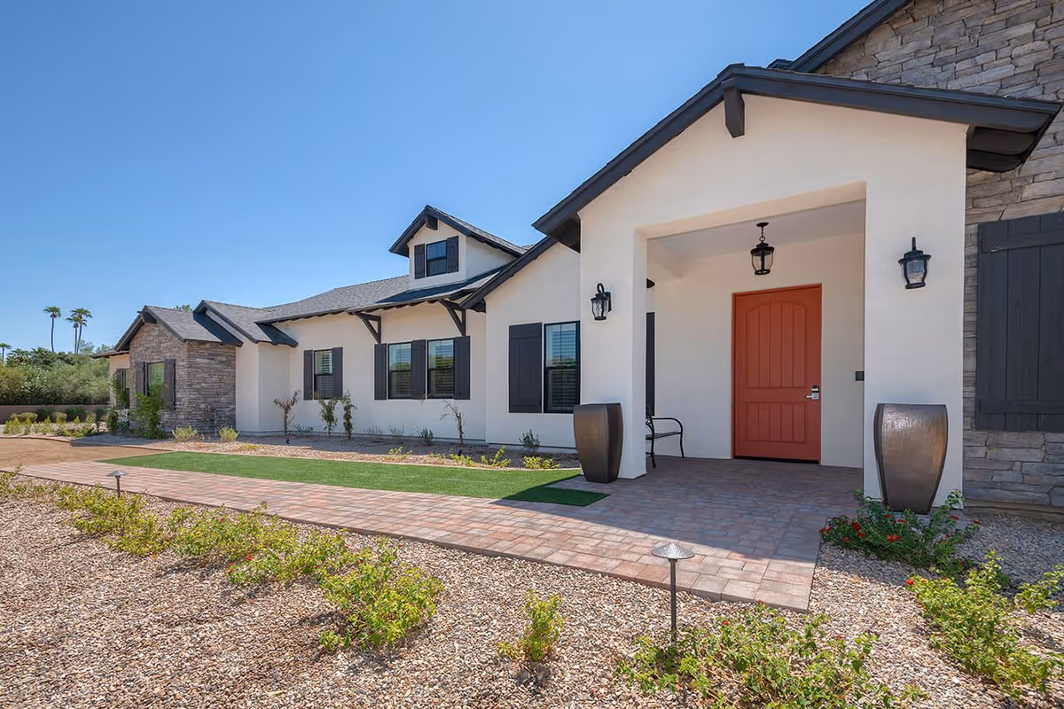 Exterior view of a single-story building with a beige stucco finish and stone accents, featuring a covered entrance with a red door, black shutters on the windows, two large decorative planters, and a paved walkway surrounded by gravel and small plants under a clear blue sky.