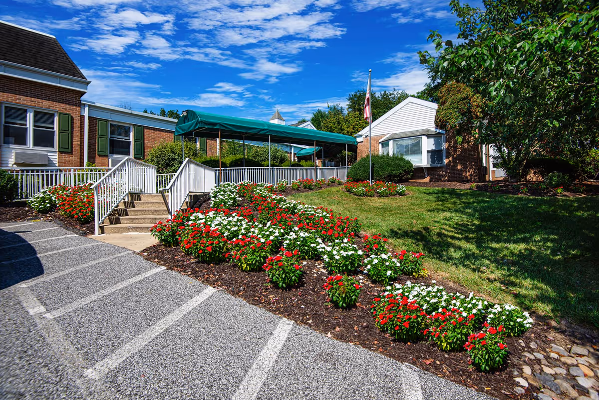 Exterior view of Brinton Manor Nursing and Rehabilitation Center showing a brick building with green shutters, a ramp and stairs leading to the entrance covered by a green canopy, landscaped flower beds with red and white flowers, a flagpole, and a clear blue sky with scattered clouds.
