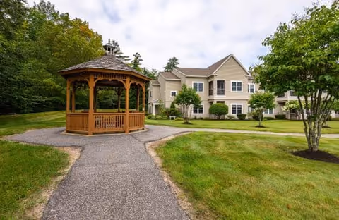 A wooden gazebo with a shingled roof sits on a paved pathway surrounded by green grass and trees. In the background, there is a two-story beige residential building with multiple windows and balconies under a partly cloudy sky.