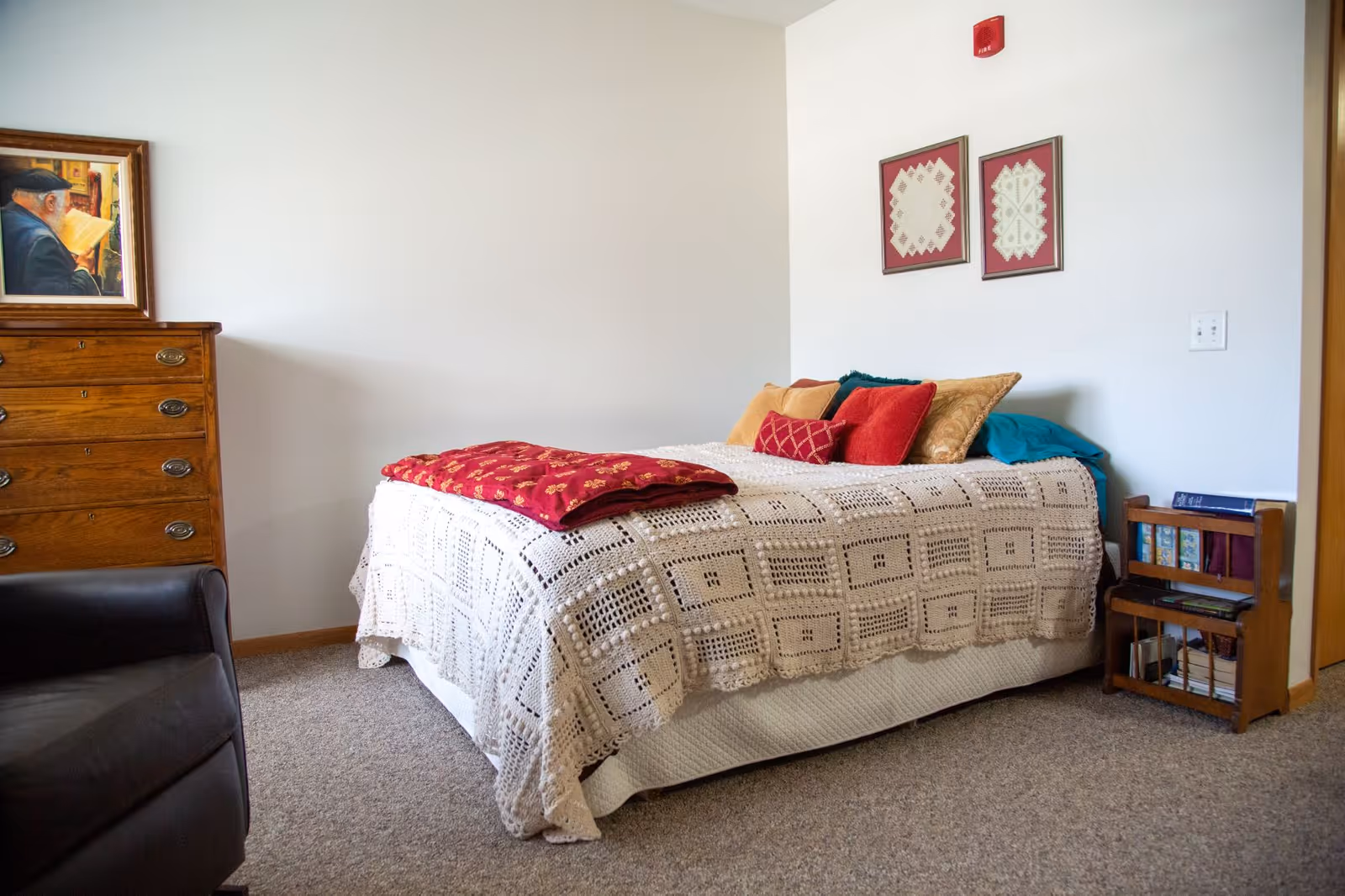 A cozy bedroom with a single bed covered in a white crocheted blanket and several colorful pillows. A red patterned throw is folded at the foot of the bed. To the left is a wooden dresser with a framed painting above it, and a black armchair is partially visible. On the right side, there is a small wooden bookshelf with books and a white wall with two framed artworks and a red fire alarm.