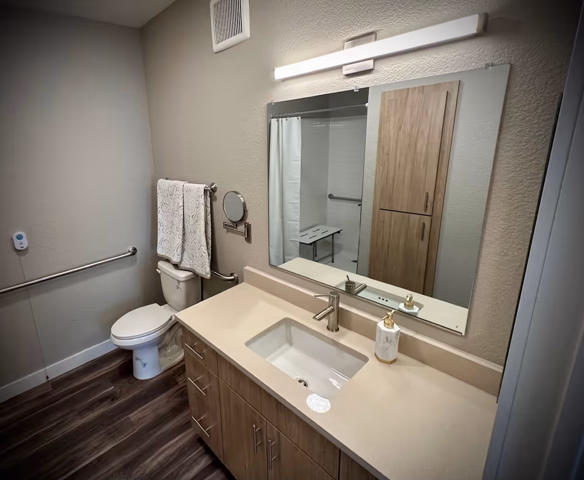 A clean and modern bathroom featuring a toilet with two white towels hanging on a bar above it, a large mirror above a beige countertop with a sink and soap dispenser, wooden cabinets below the sink, a wall-mounted light above the mirror, and a shower area with a white curtain and a foldable shower seat visible in the reflection.