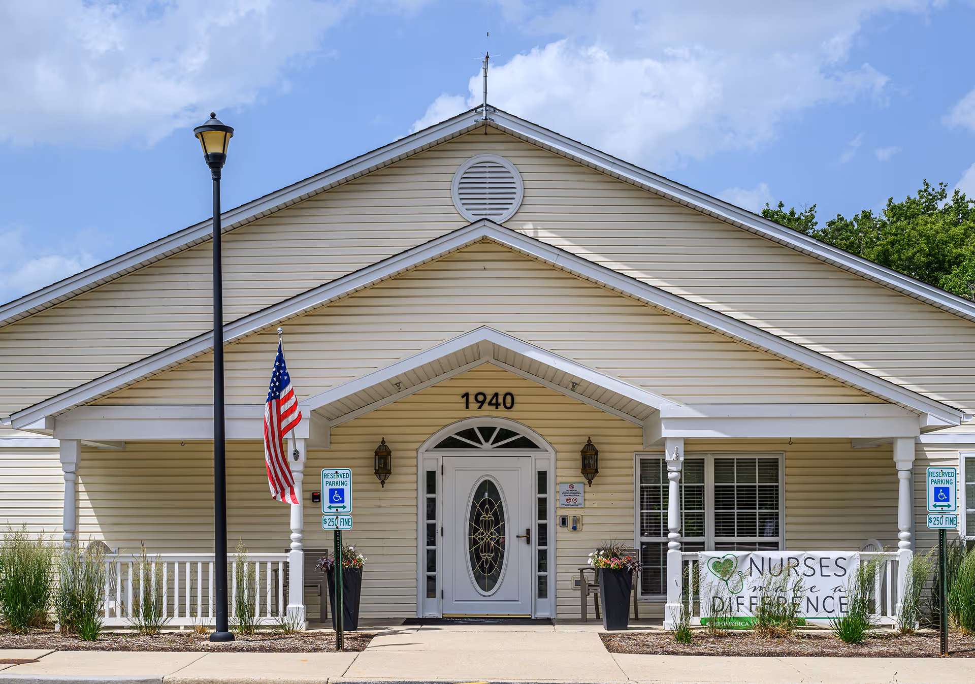 Front entrance of a beige single-story building numbered 1940 with a porch, American flag, handicap parking signs, and a banner reading 'Nurses make a difference'.
