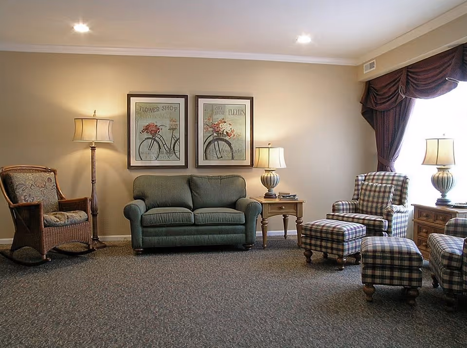 A cozy living room with a green loveseat, a wicker armchair with floral cushions, two plaid armchairs with matching ottomans, two wooden side tables each with a lamp, and two framed floral bicycle prints on the beige wall. The room has carpeted flooring and a window with dark drapes.