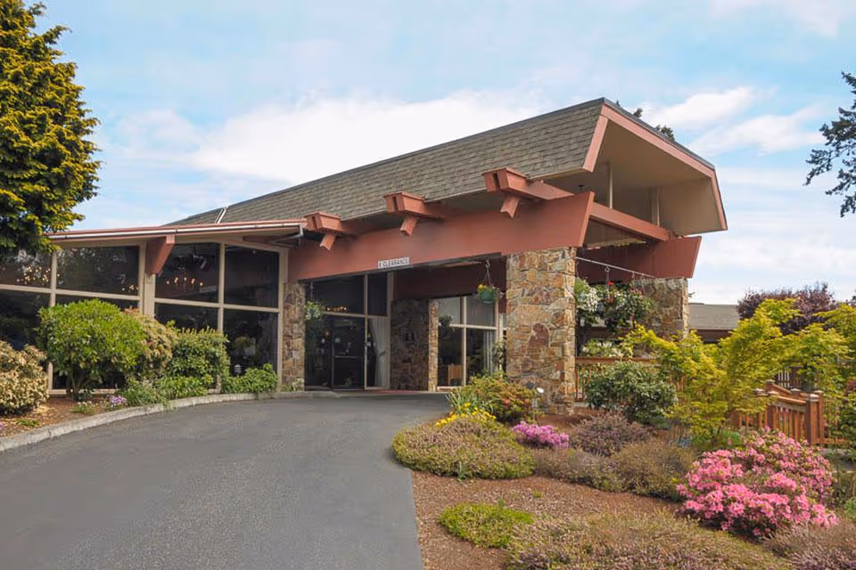Covered entrance and porte-cochère of a single-story building with stone columns, large windows and landscaped flowering shrubs.