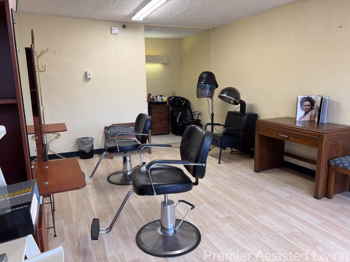 Interior view of a small salon area with two black salon chairs in front of mirrors, a hair washing station with a black chair and sink in the back, a hair dryer, and a wooden desk with a photo of a woman on it. The floor is light wood, and the walls are beige.