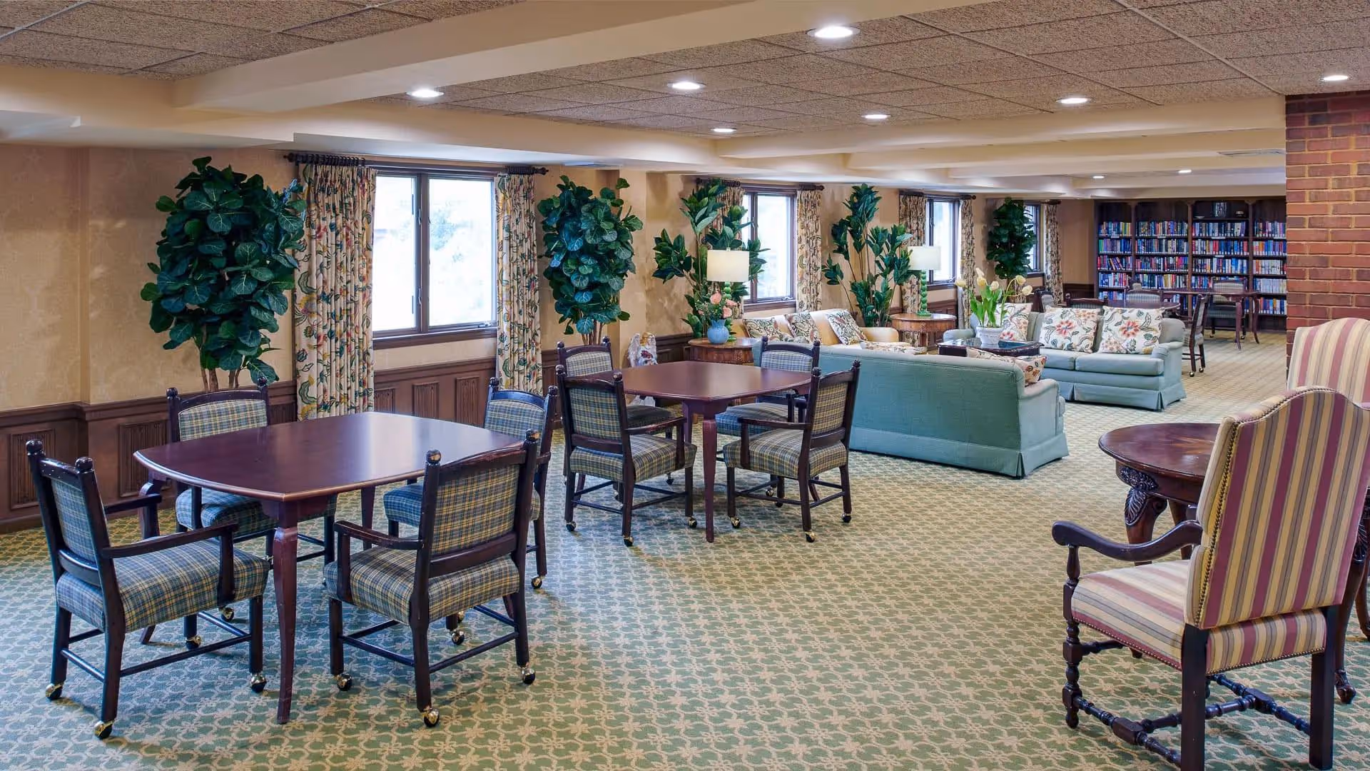 Spacious senior living common room with tables and chairs in the foreground, sofas and bookshelves in the background, and potted plants by the windows.