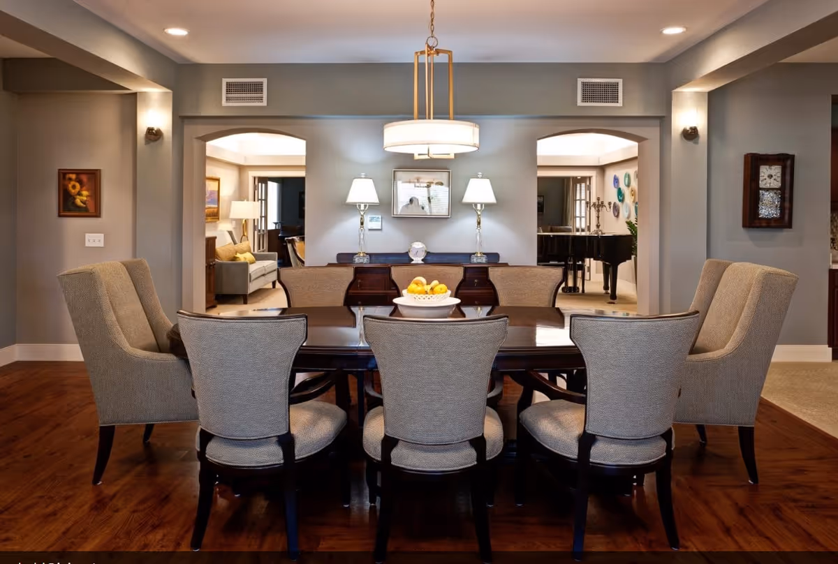 A formal dining room with a dark wooden table surrounded by eight upholstered chairs. A bowl of fruit is placed in the center of the table. The room features wooden flooring, gray walls, and a modern chandelier hanging above the table. Two table lamps sit on a sideboard against the far wall, with framed artwork above it. Two archways lead to adjacent rooms, one with a sofa and lamp, and the other with a grand piano and decorative plates on the wall.
