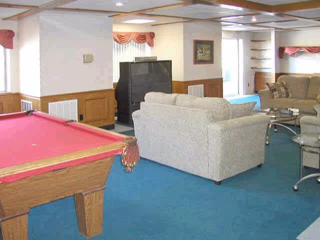 A common area with a red pool table in the foreground, beige sofas arranged around a glass coffee table, a television set, and windows with pink valances. The room has blue carpet and wood paneling on the lower half of the walls.