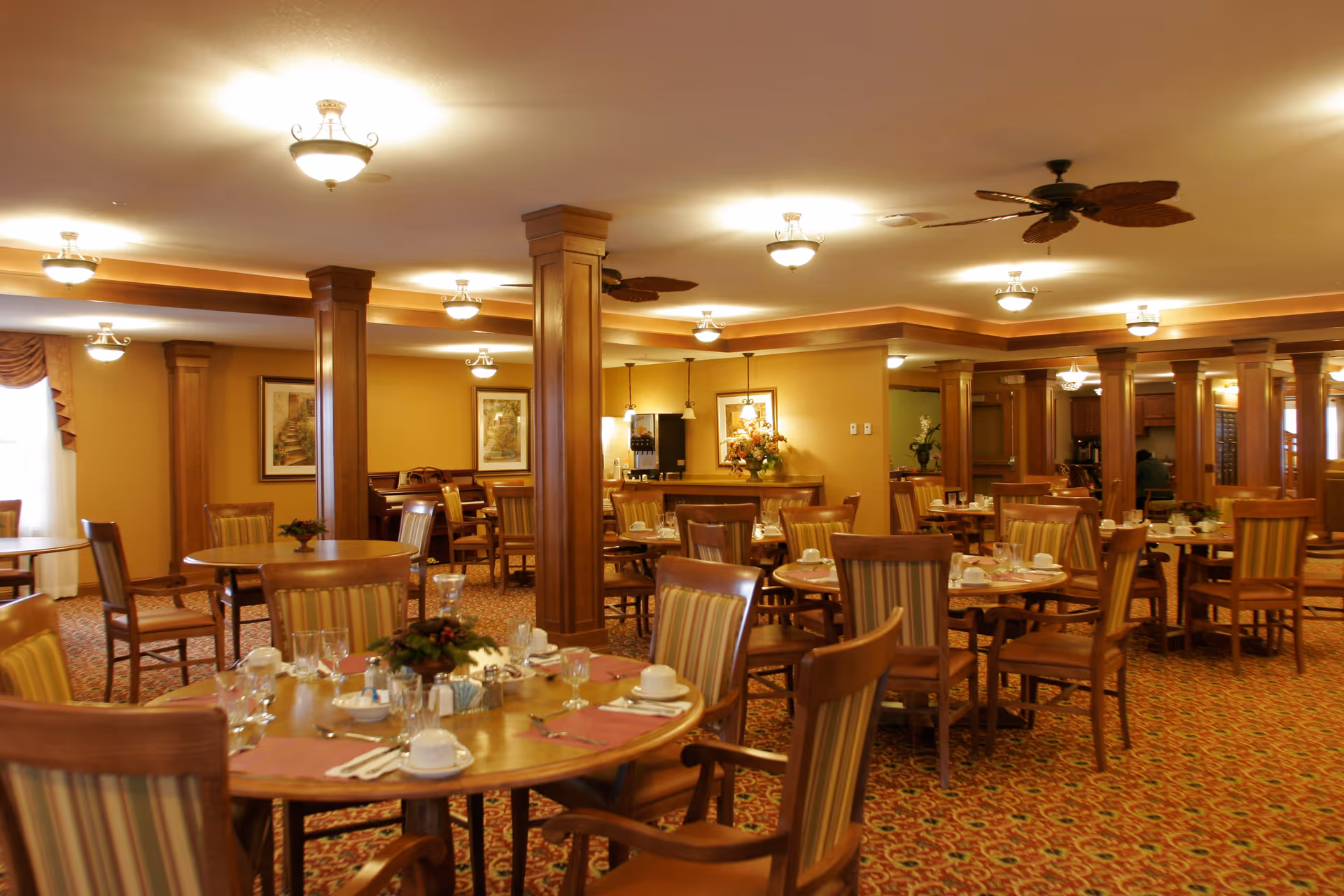 A warm and inviting dining room with multiple round wooden tables and striped cushioned chairs. Each table is set with white cups, glasses, silverware, and napkins. The room features wooden columns, framed artwork on the walls, ceiling lights, and ceiling fans. The carpet has a patterned design, and there is a beverage station in the background.