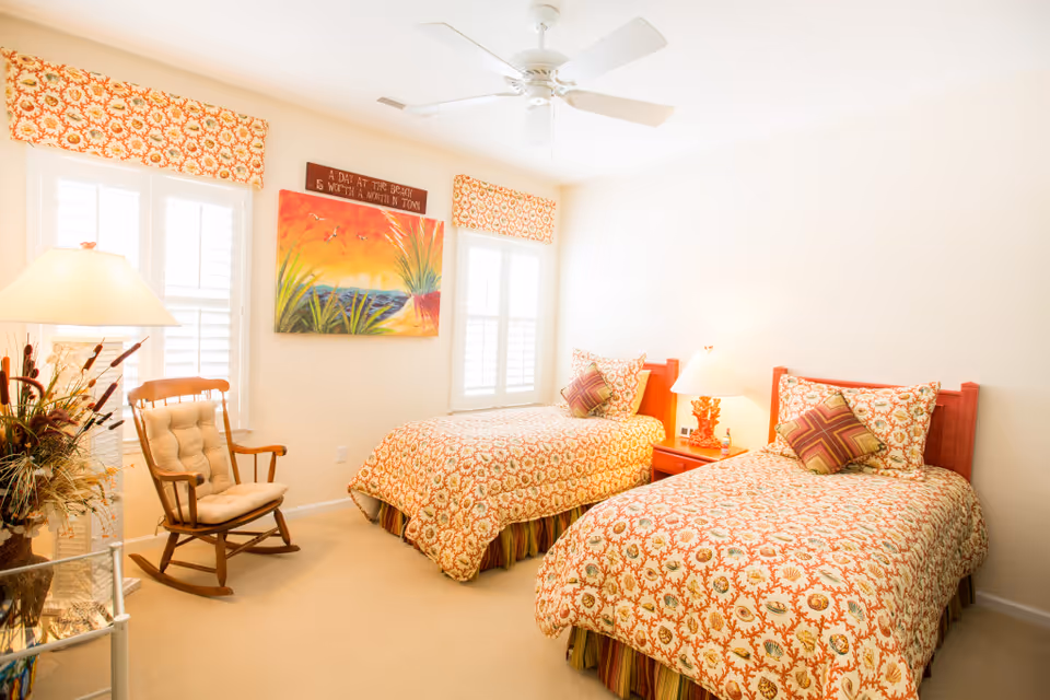 A bright bedroom with two twin beds featuring matching orange and white patterned bedspreads and pillows. There is a wooden rocking chair with a cushion near two windows with white shutters and orange patterned valances. A colorful painting of a beach scene hangs on the wall between the windows, with a sign above it that reads 'A day at the beach is worth a month in town.' A ceiling fan is mounted on the white ceiling, and a bedside table with a lamp is positioned between the beds.