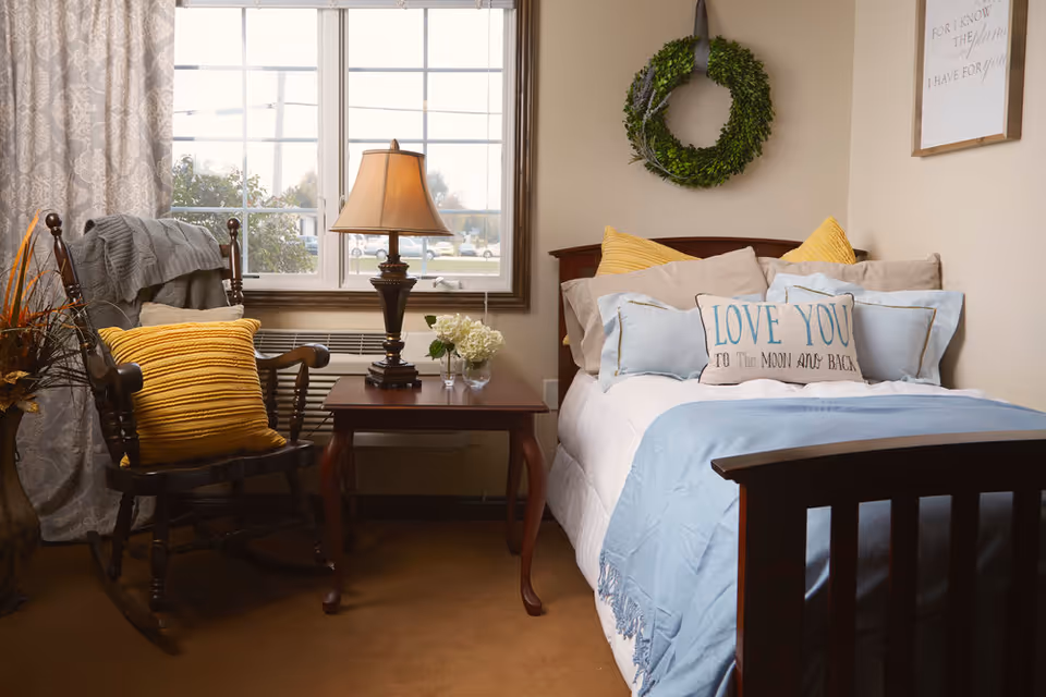 A cozy bedroom with a wooden bed dressed in light blue and beige bedding, including a decorative pillow that says 'LOVE YOU TO THE MOON AND BACK.' A green wreath hangs on the wall above the bed. Next to the bed is a wooden side table with a lamp and a small vase of white flowers. A wooden rocking chair with a yellow pillow and a gray blanket is positioned near a window with patterned curtains.