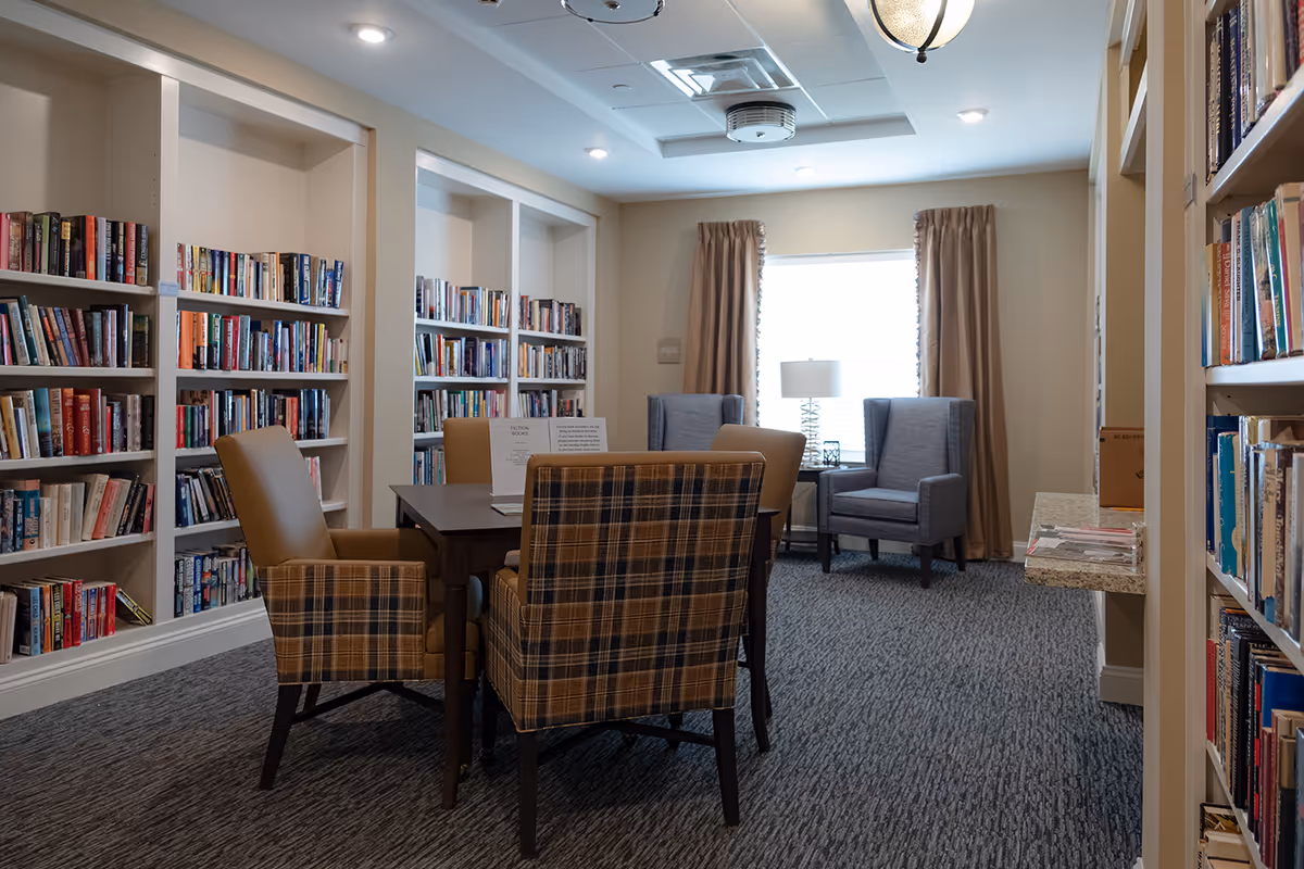 A cozy reading room with bookshelves filled with books lining the walls. There is a wooden table surrounded by four chairs, two of which have a plaid pattern. In the background, two gray armchairs are positioned near a window with beige curtains and a table lamp between them. The room has carpeted flooring and soft lighting from ceiling fixtures.
