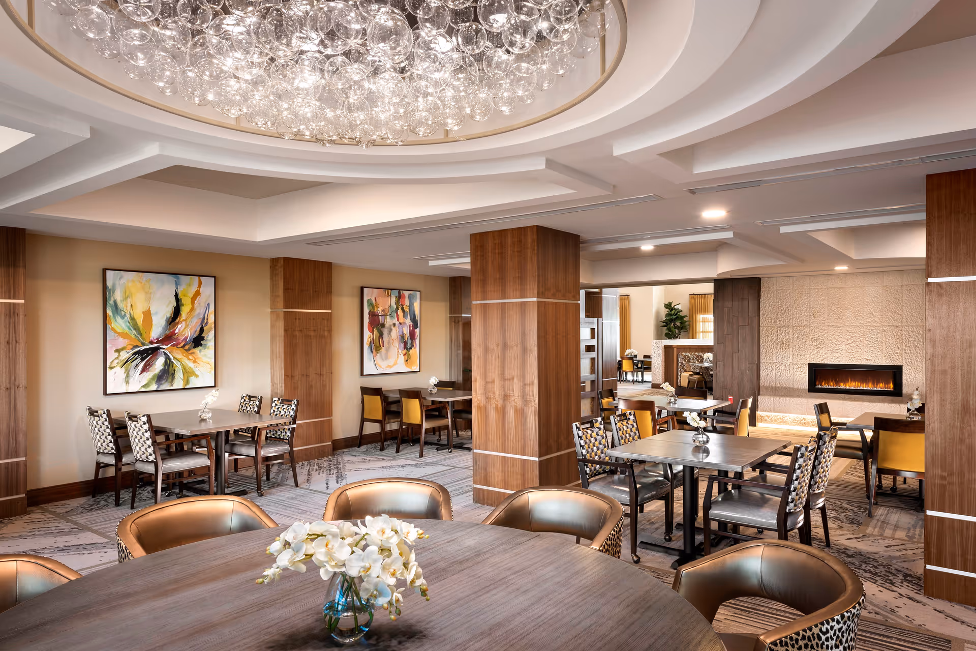 Elegant communal dining and lounge area with tables, chairs, a large chandelier, artwork on the walls, and a fireplace.