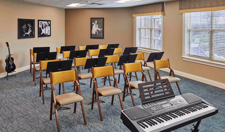 A music room with several rows of chairs each equipped with a black music stand. There is a keyboard on a stand in the foreground and an acoustic guitar resting against the wall on the left. The room has beige walls with three black and white framed photos and two large windows with blinds letting in natural light.