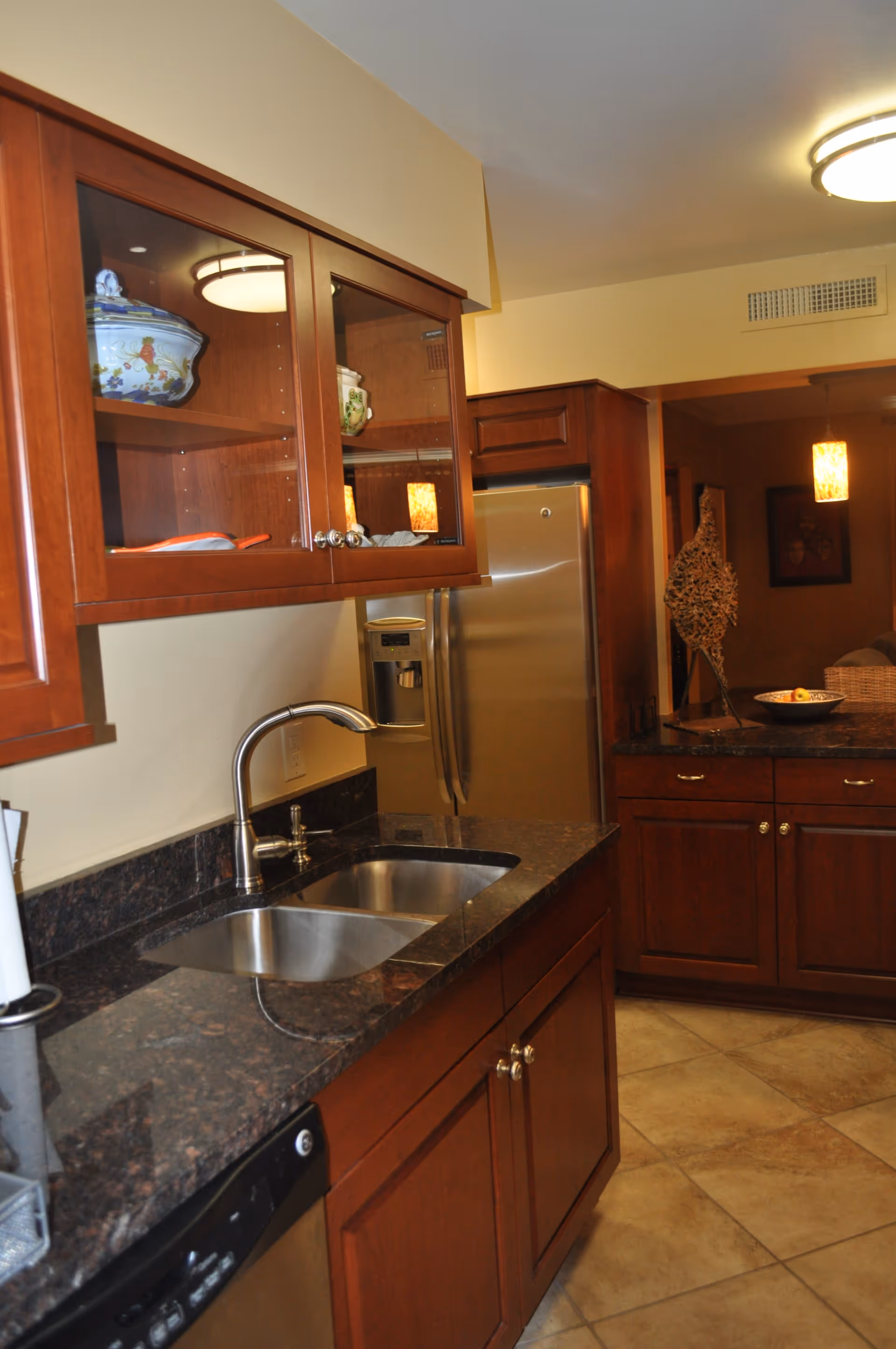 Kitchen area with dark granite countertops, wooden cabinets with glass doors displaying decorative items, a stainless steel double sink with a modern faucet, a stainless steel refrigerator, and tiled flooring. The kitchen opens into a dimly lit living area with a decorative sculpture and a bowl of fruit on the counter.