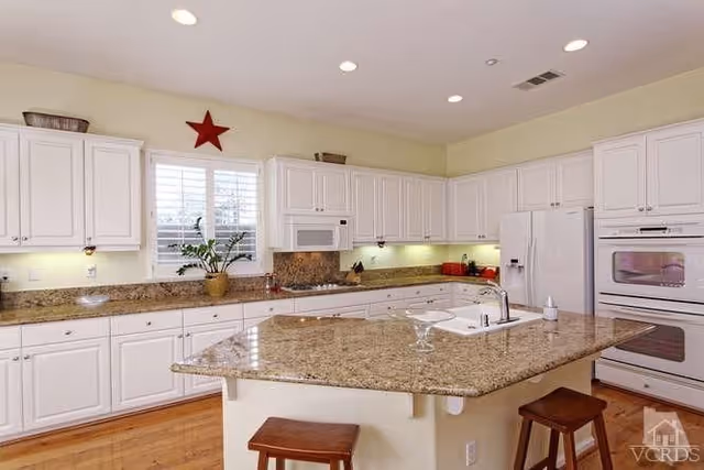 Bright kitchen with a large granite island, white cabinets, stainless appliances, and two stools.