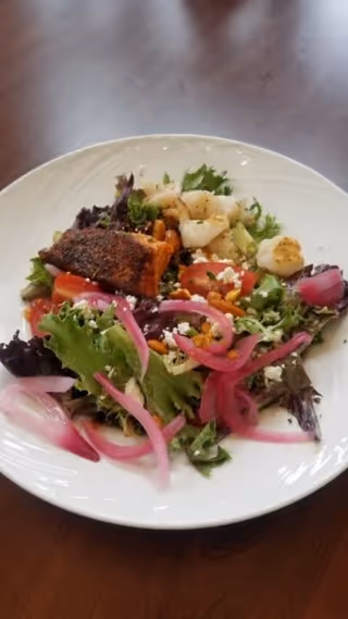 A white plate with a fresh salad consisting of mixed greens, pickled red onions, cherry tomatoes, crumbled cheese, pine nuts, and a piece of seasoned cooked salmon on top.