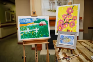 Three colorful paintings displayed indoors: one on an easel depicting a rural landscape with a red barn and white fence, another on an easel showing pink flowers on a yellow background, and a smaller framed painting of a bird on a table with paintbrushes nearby.