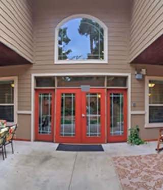 Entrance area of a building with red double doors featuring glass panels. Above the doors is a large arched window. The exterior walls are beige with horizontal siding. There are chairs and a table on the left side and a patterned rug on the right side of the concrete floor.