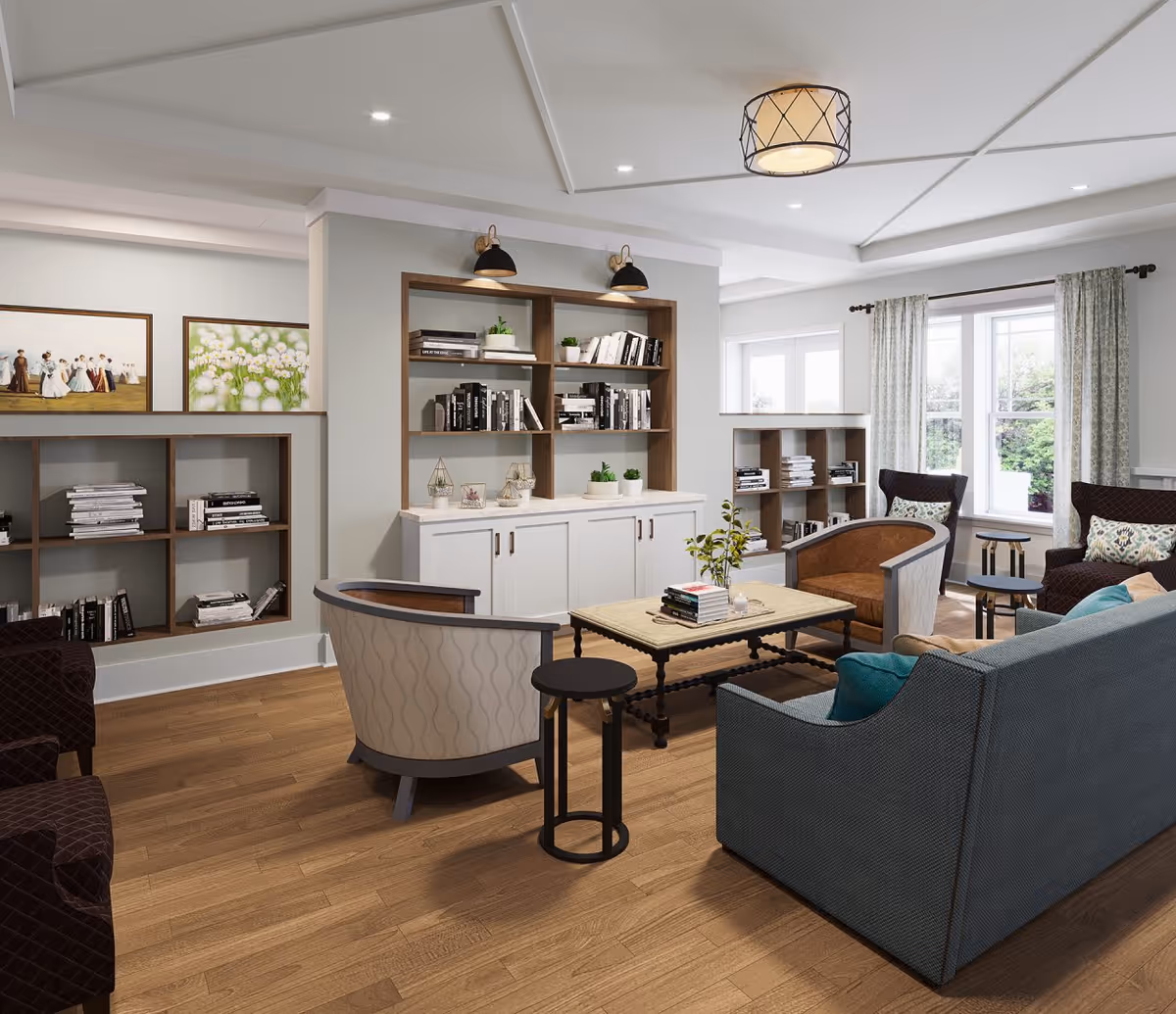A cozy living room area with wooden flooring, a blue sofa, two armchairs, a coffee table with books, built-in bookshelves filled with books and decorative plants, large windows with curtains letting in natural light, and framed artwork on the walls.