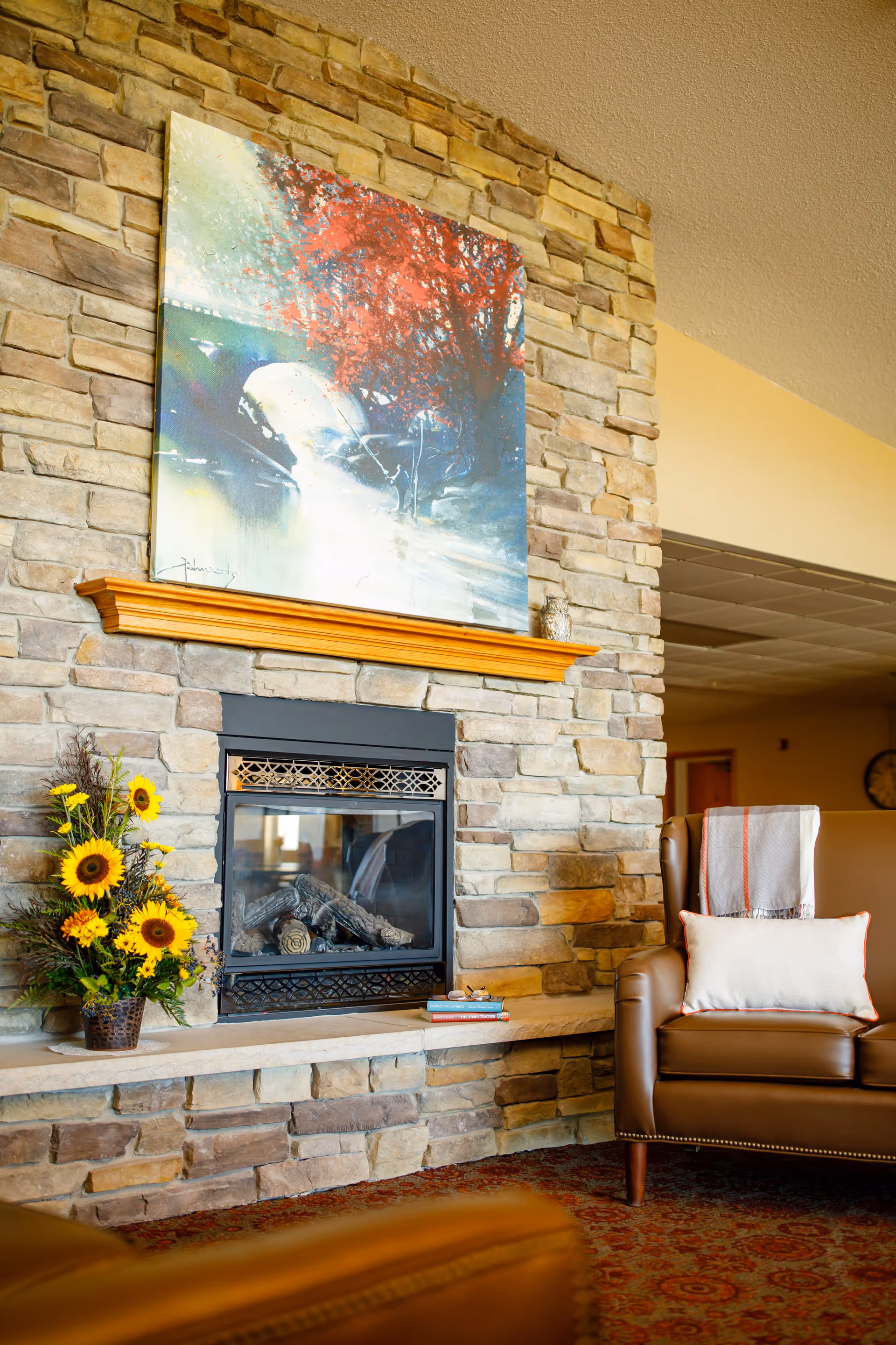 Cozy living room area with a stone fireplace featuring a wooden mantel. Above the mantel hangs a colorful painting of a bridge and red trees. To the left of the fireplace is a vase with sunflowers and other flowers. To the right is a brown leather armchair with a white pillow and a folded blanket draped over the back. The room has warm lighting and a patterned carpet.