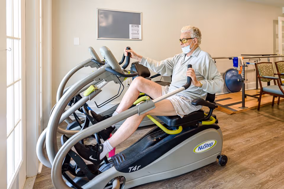 An elderly person wearing a face mask and glasses exercises on a NuStep recumbent cross trainer machine in a well-lit room with wooden flooring and chairs in the background.