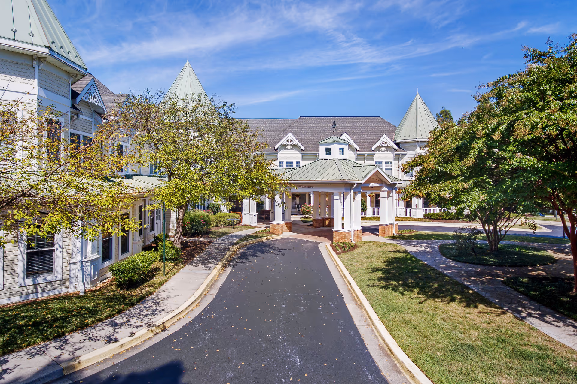 Front entrance and driveway of a large white senior living building with a covered porte-cochère and landscaped trees.