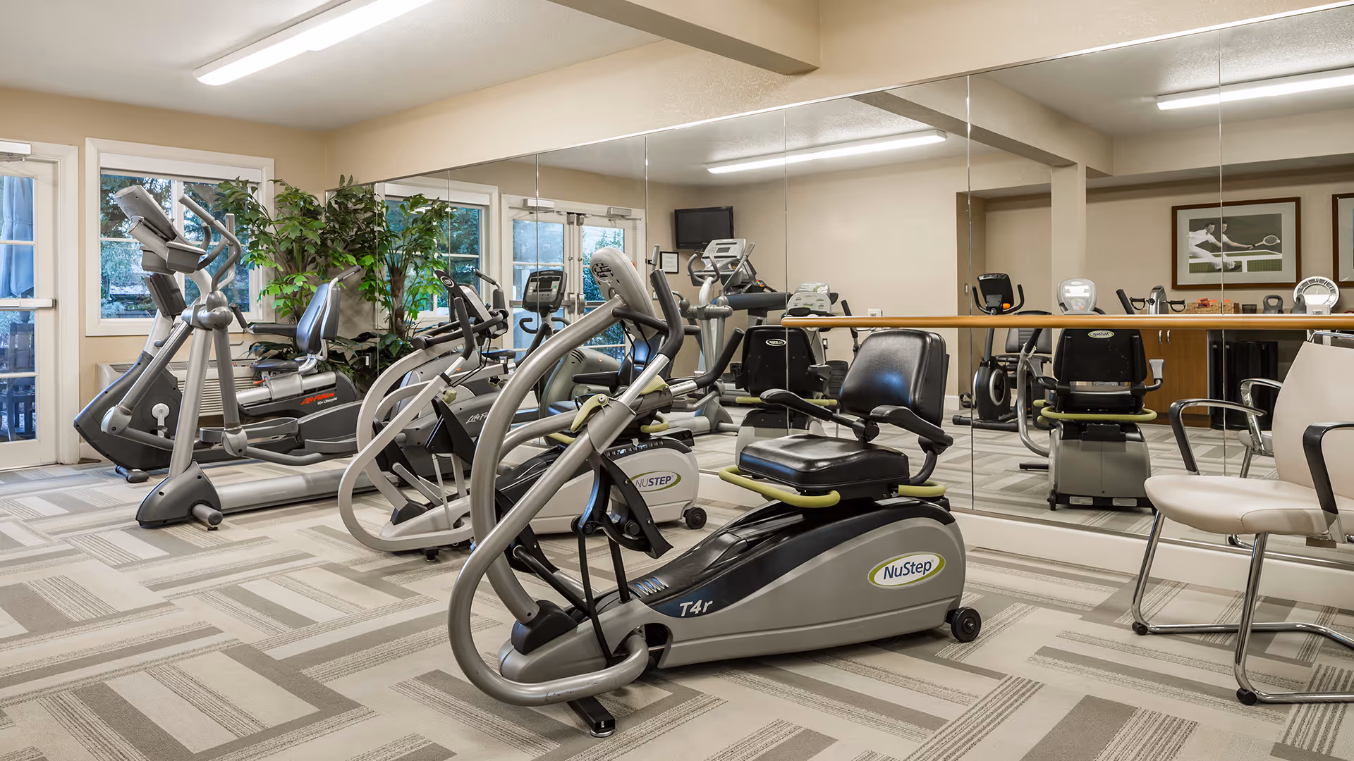 A well-lit fitness room with various exercise equipment including NuStep recumbent cross trainers, treadmills, and stationary bikes. The room has large mirrors on one wall, windows letting in natural light, and a patterned carpeted floor. There are also a few chairs and framed pictures on the wall.