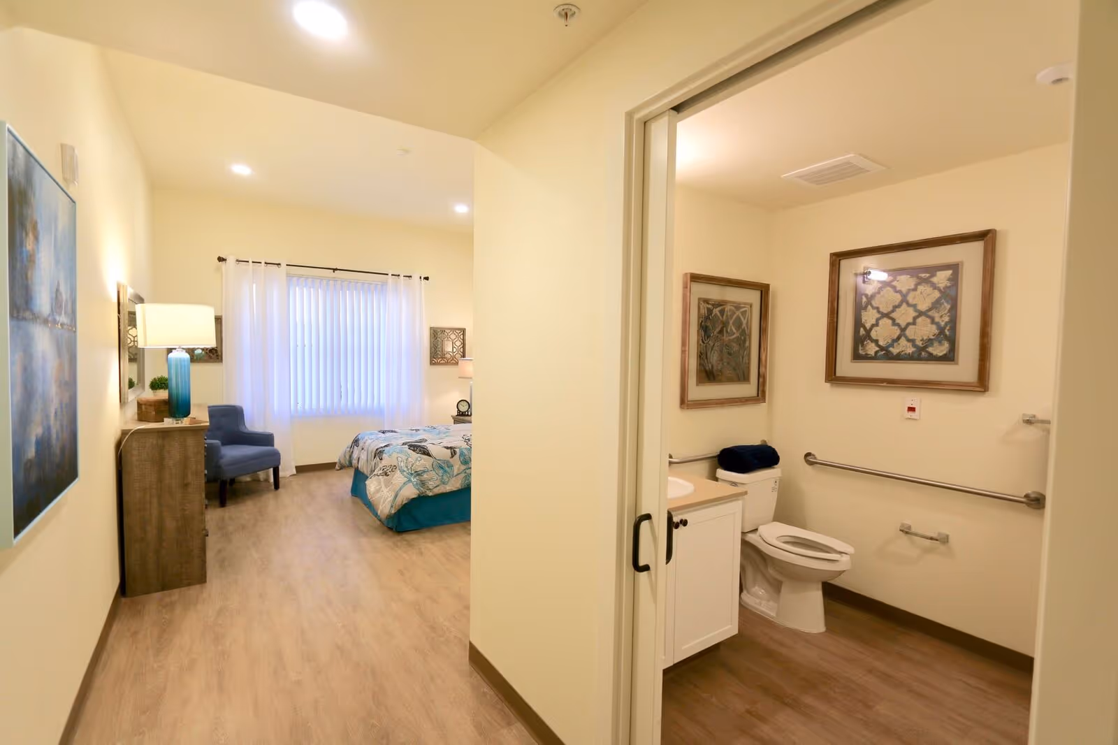 View of a senior living facility room showing a bedroom area with a bed, blue chair, dresser with a lamp, and artwork on the walls. To the right, there is an open door leading to an accessible bathroom with a toilet, sink, grab bars, and framed artwork.