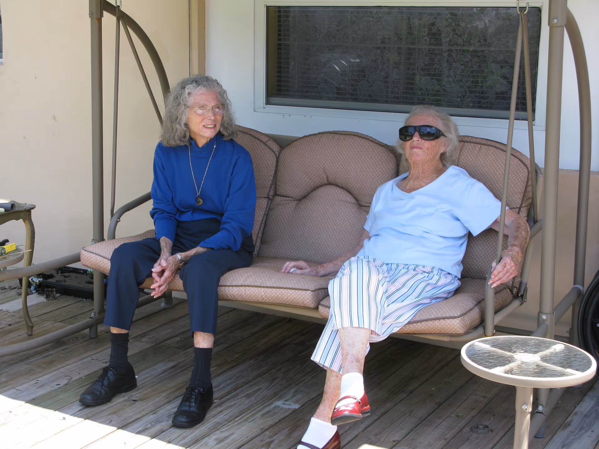 Two elderly women sitting on a cushioned outdoor swing on a wooden deck. One woman is wearing a blue sweater and dark pants, while the other is wearing a white shirt, striped pants, and sunglasses. There is a small round glass table next to the swing and a window behind them.