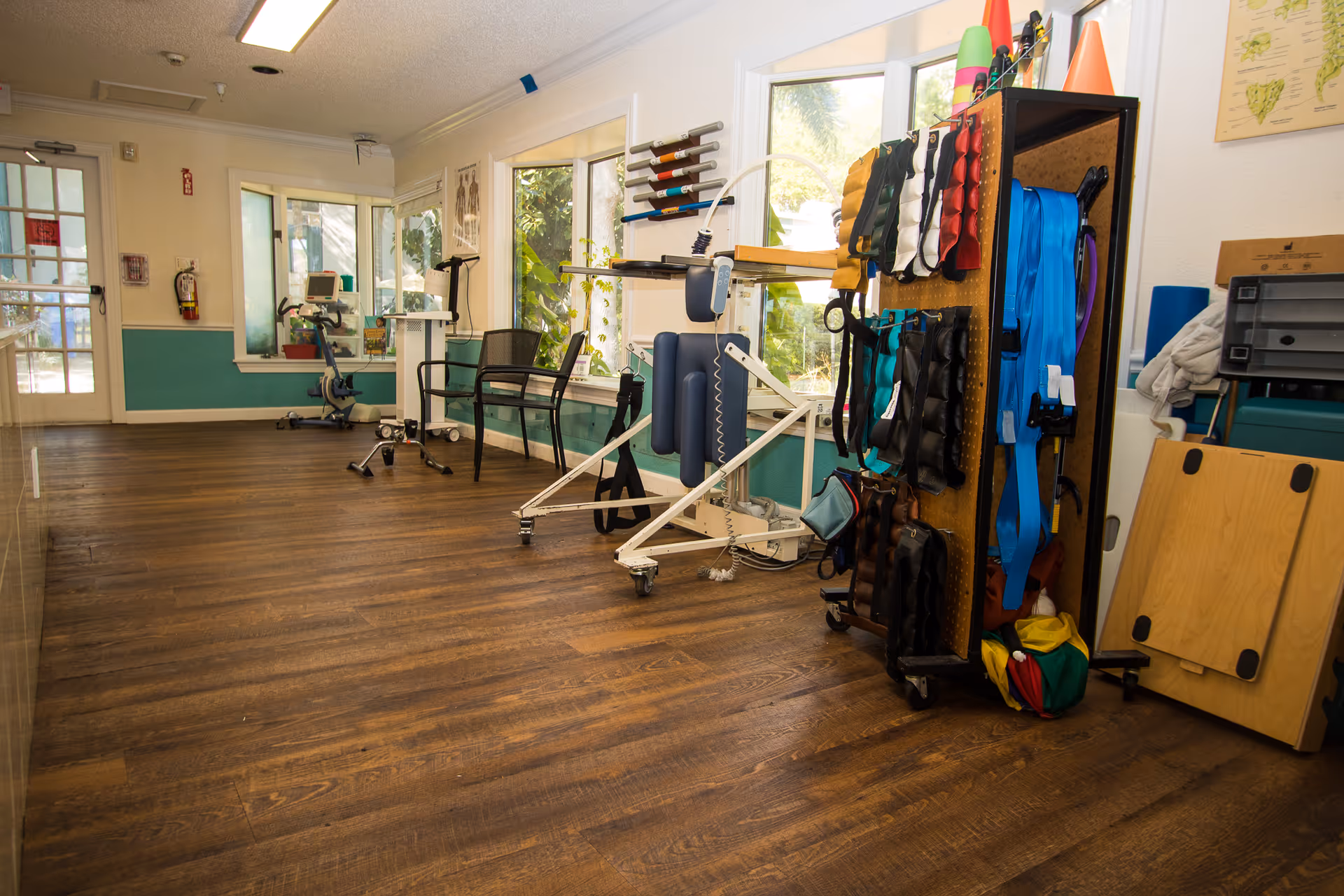 Rehabilitation therapy room with exercise equipment, a patient lift, chairs, and a rack of straps and therapy supplies on wood flooring.