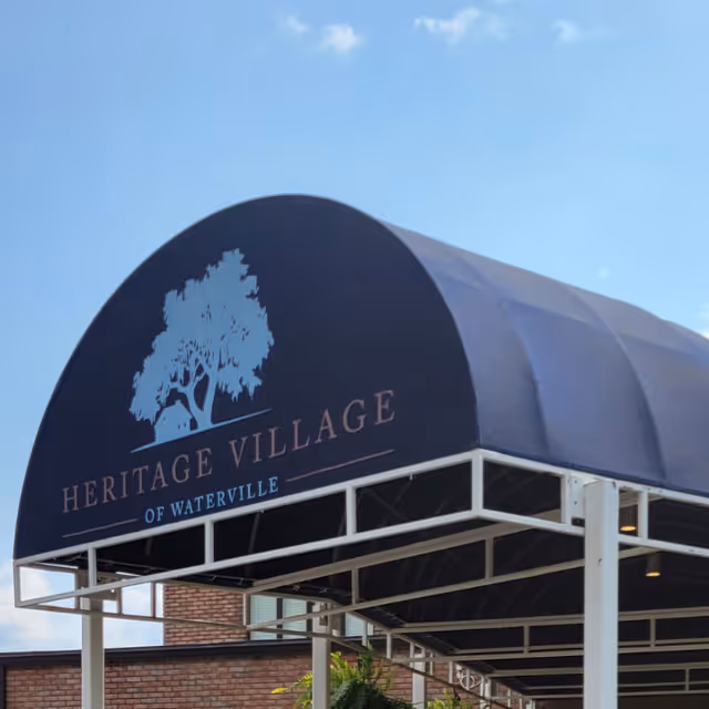 A blue canopy with white frame displaying the text 'Heritage Village of Waterville' and a tree logo above the text, set against a partly cloudy blue sky.