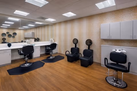 Interior salon room with styling chairs, large mirror, hair dryers, sinks and cabinetry on wood-look flooring.