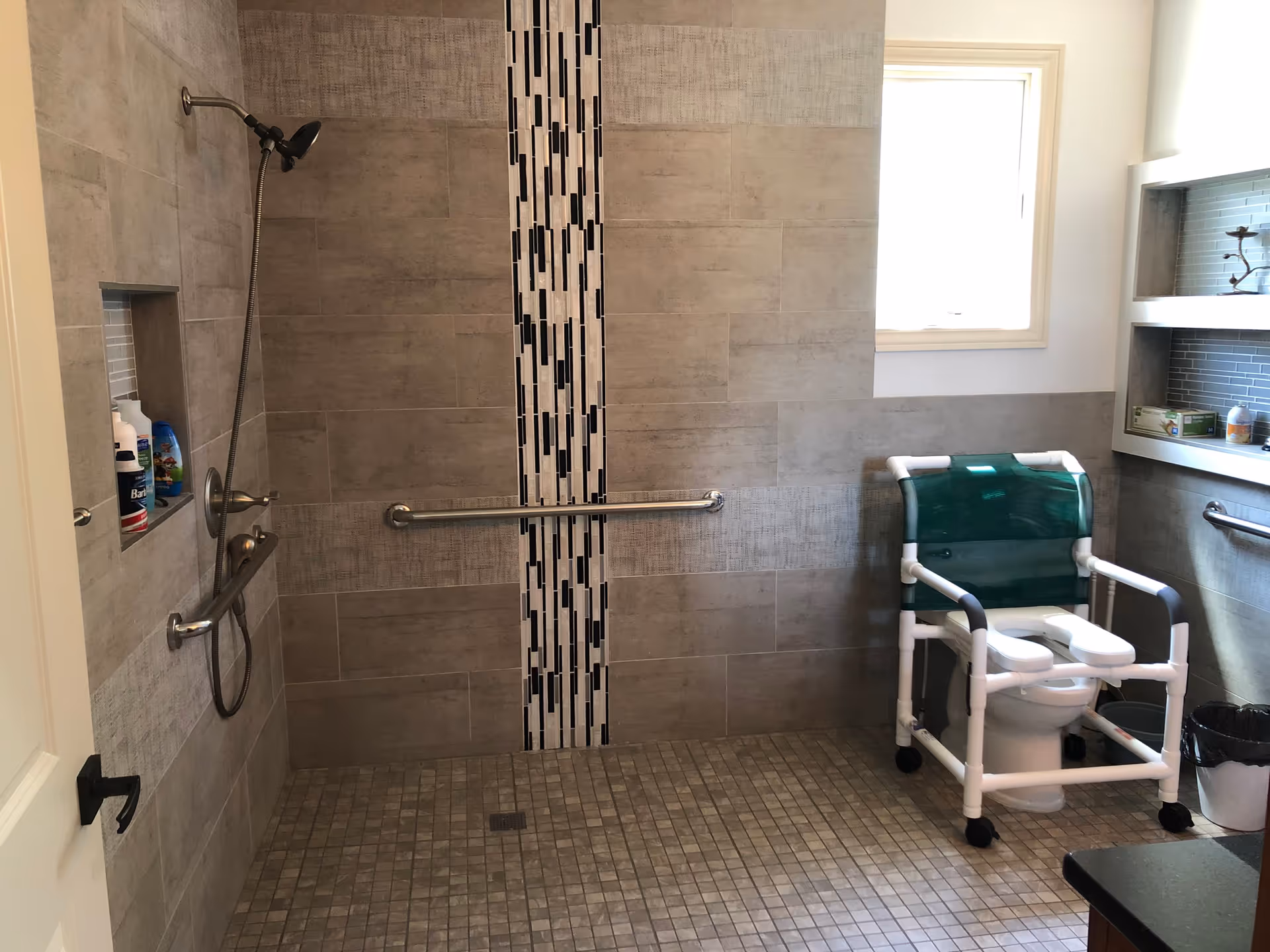 Tiled accessible bathroom with a walk-in shower, horizontal grab bar, handheld showerhead, and a mobile commode chair by a window and shelving.