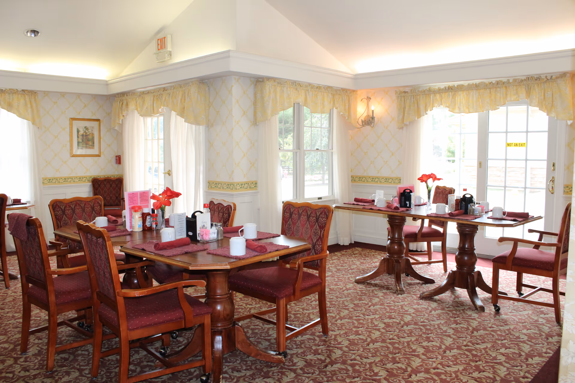 A dining room with two wooden tables set with red placemats, white mugs, napkins, and condiments. The room has patterned carpet, wallpaper with a yellow diamond pattern, and windows with sheer white curtains and yellow valances. There are wooden chairs with red upholstered seats and backs around the tables. The room is well-lit with natural light coming through the windows and a wall sconce light fixture.