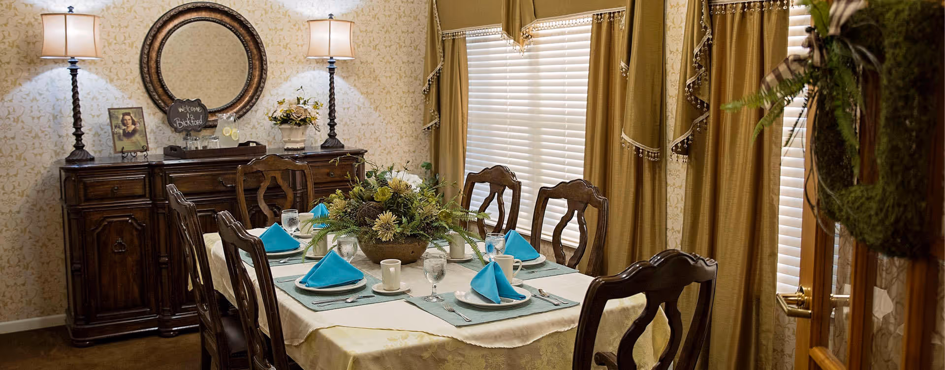 A formal dining room with a rectangular table set for six people. The table is covered with a cream tablecloth and has blue napkins folded on white plates, along with glasses and silverware. A floral centerpiece decorates the middle of the table. Behind the table is a wooden sideboard with two lamps, a round mirror, a framed photo, and a small chalkboard sign that says 'Welcome to Bickford.' The room has patterned wallpaper and large windows with gold curtains.