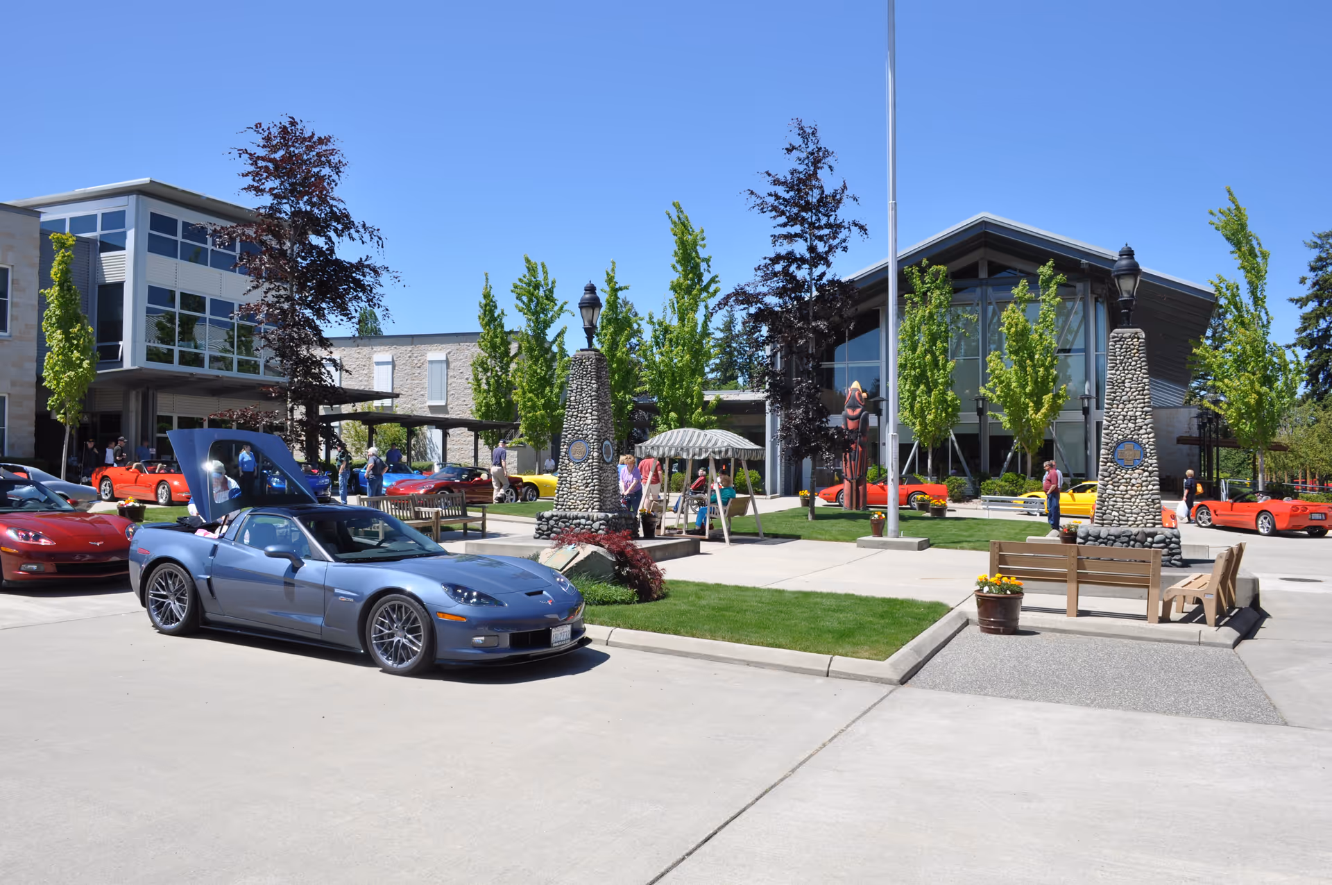 Front courtyard of the Washington Veterans Home with a modern building entrance, stone pillars, benches, trees, several parked sports cars, and people under a clear blue sky.