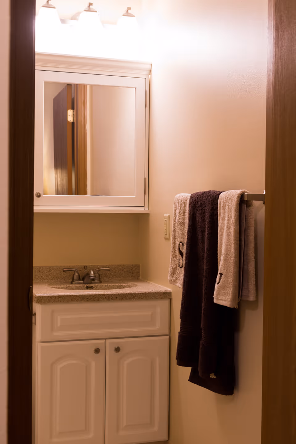 Small bathroom vanity with a granite countertop, a sink with a faucet, a white cabinet below, a wall-mounted mirror cabinet above, and three towels hanging on a towel rack on the right wall.