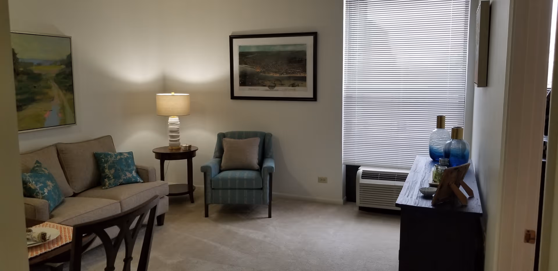 A cozy living room with a beige sofa adorned with blue patterned pillows, a blue armchair with a gray cushion, a round wooden side table with a lamp, a framed picture on the wall, a window with closed blinds, and a wooden cabinet with decorative blue glass vases and other small items.