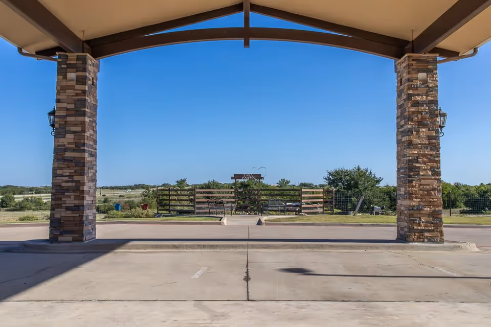 View from under a covered entrance with stone pillars looking out onto a fenced grassy area with benches and a sign that reads 'The Farm'. Clear blue sky and open landscape in the background.