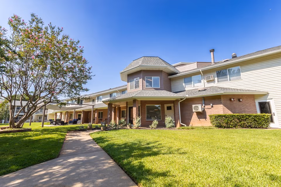 Exterior view of a two-story senior living facility building with a covered porch area, surrounded by a well-maintained lawn and a tree with pink flowers under a clear blue sky.