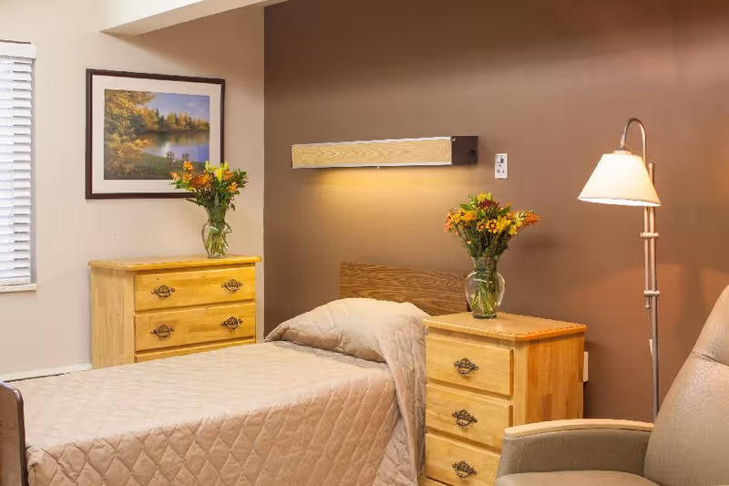 Single bed in a cozy bedroom with wooden nightstands, vases of flowers, a floor lamp, and framed artwork.