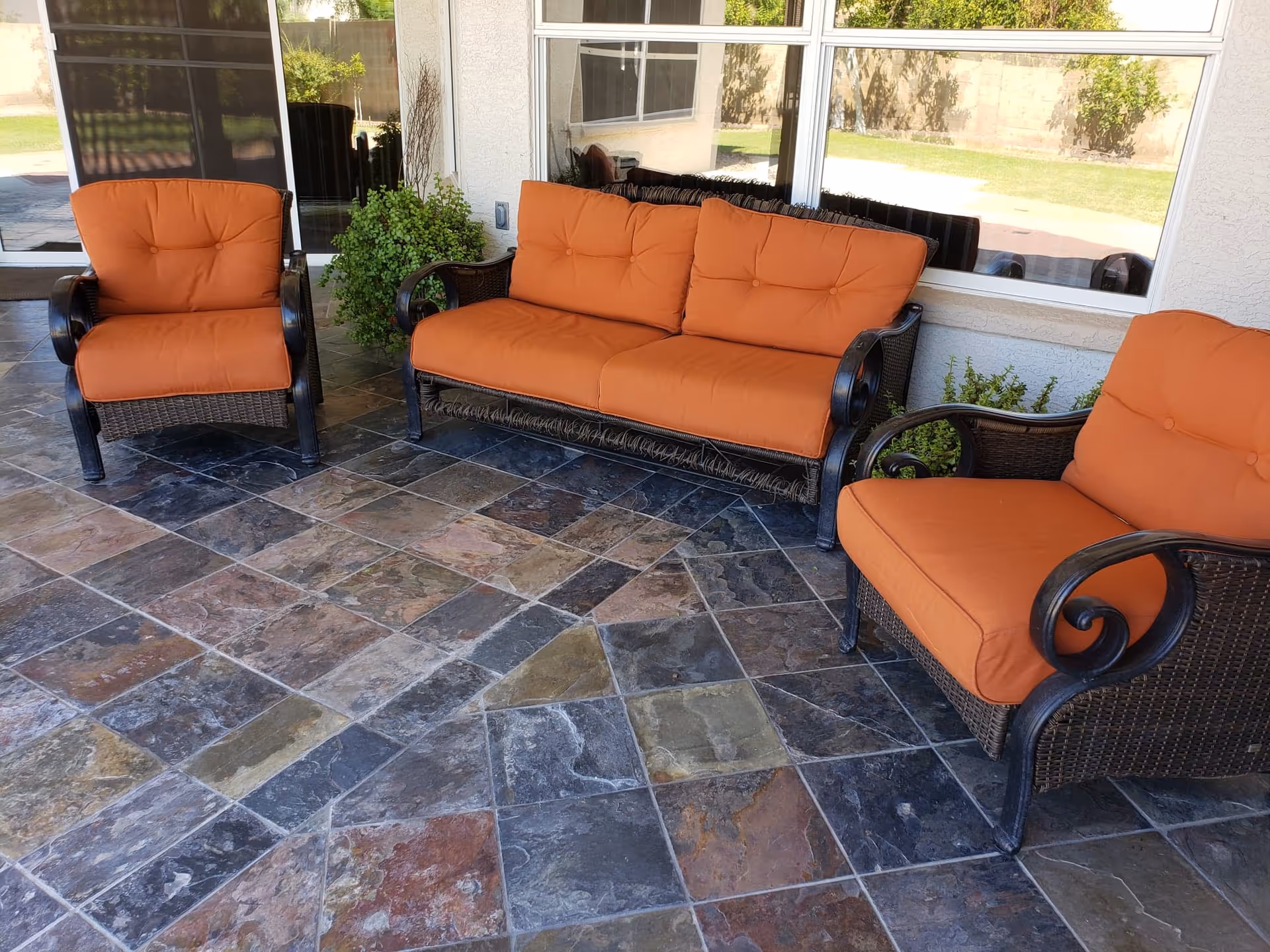 Outdoor patio area with three cushioned wicker chairs and a loveseat with orange cushions, set on a tiled floor with a window and plants in the background.