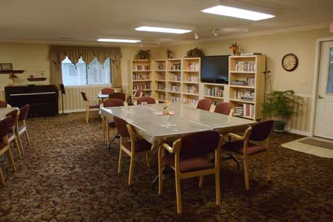 A common room with a large rectangular table covered with a plastic tablecloth surrounded by chairs. Bookshelves filled with books and a television are against the far wall. There is a piano on the left side, a window with curtains, a clock on the wall, and a potted plant near the door on the right.