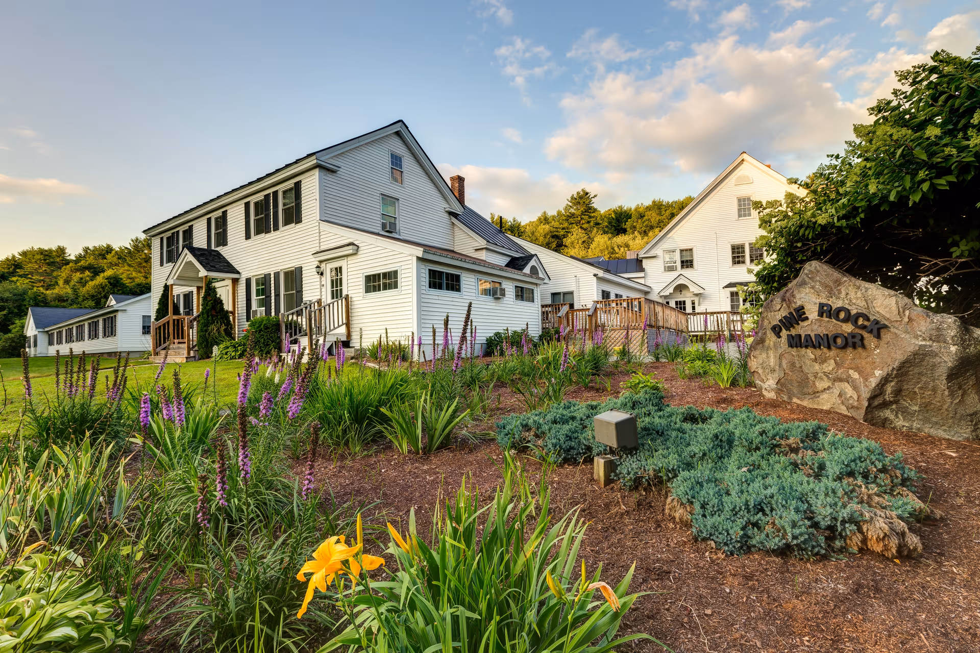 Exterior view of Pine Rock Manor, a white multi-story building surrounded by a well-maintained garden with purple and yellow flowers, green shrubs, and a large rock with the facility name on it under a partly cloudy sky.