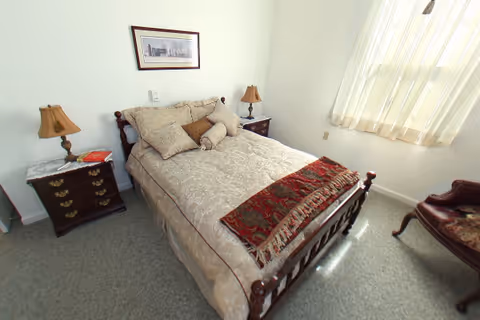 A small bedroom with a wooden bed frame and a neatly made bed with beige patterned bedding and multiple pillows. There are two dark wooden nightstands on either side of the bed, each with a lamp. A framed picture hangs on the white wall above the bed. A window with sheer white curtains allows natural light to fill the room. A wooden chair with a cushioned seat is placed near the window.