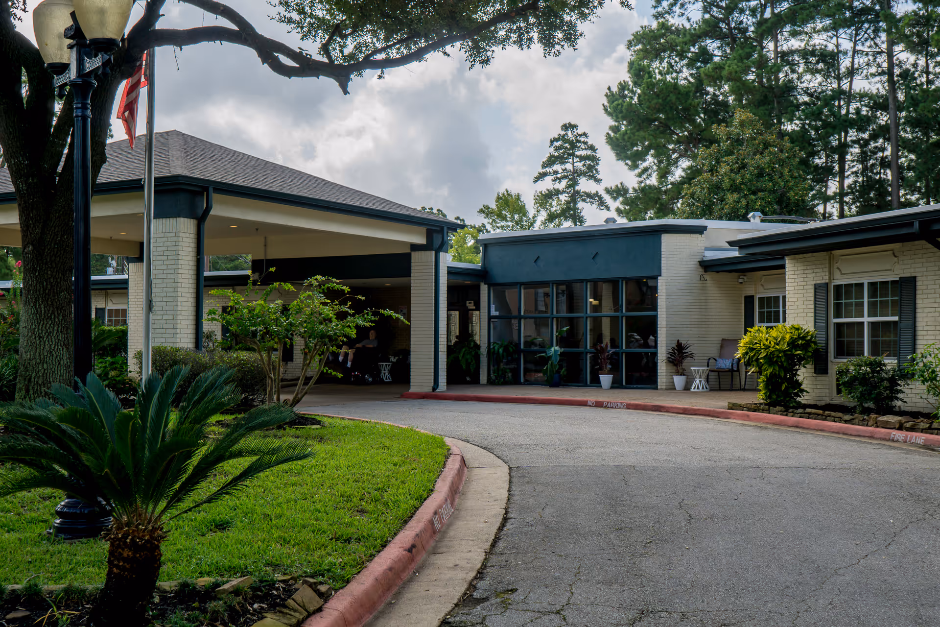 Exterior view of Huntsville Health Care Center showing the entrance with a covered drop-off area, surrounded by greenery including trees and shrubs, with a paved driveway and a lamppost with an American flag.