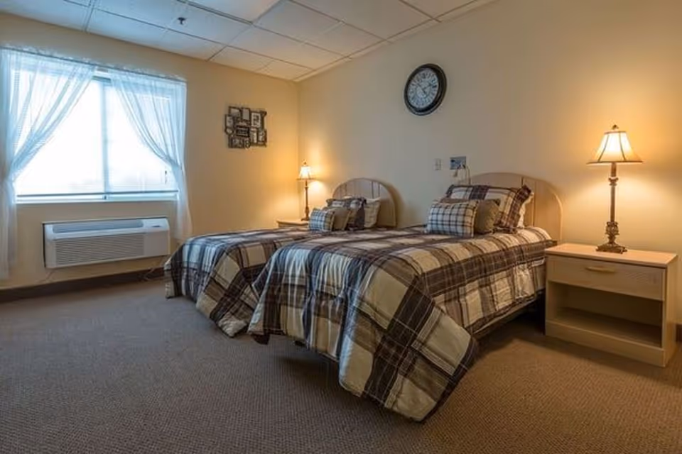 Twin beds with plaid bedding in a softly lit bedroom with nightstands, lamps, a window with curtains, and a wall clock.