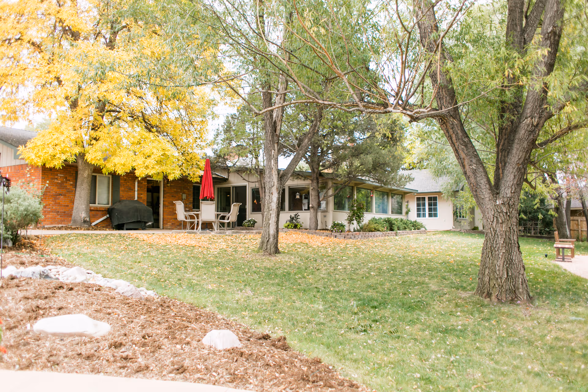 Front exterior of a single-story assisted living building with a patio, red umbrella, and trees in a grassy yard.