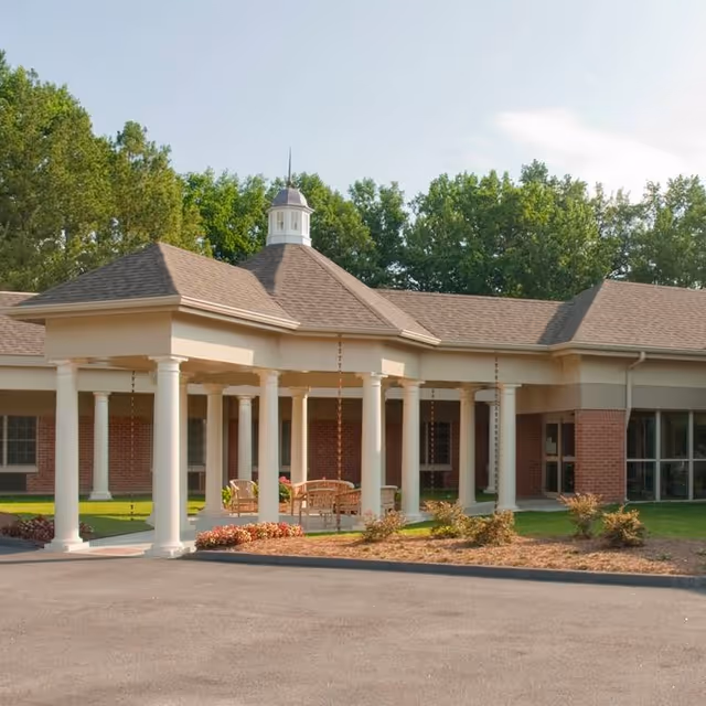Exterior view of a single-story brick building with a covered entrance supported by white columns, surrounded by greenery and trees in the background.